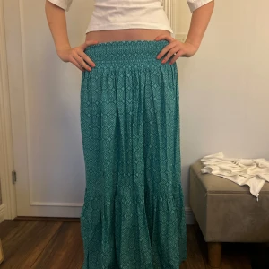 Turkos mönstrad långkjol - Säljer en turkos långkjol med geometriskt mönster och smock i midjan. Kjolen är luftig och har en lätt volang nertill, perfekt för dig som gillar boho-vibe och vill ha något bekvämt men ändå snyggt. Passar till både sneakers och sandaler.