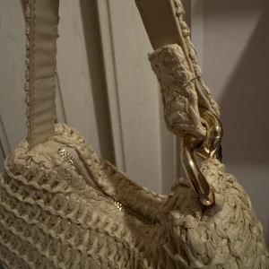 Beige axelväska i flätad syntet - Snygg beige axelväska med flätad struktur och guldfärgade metalldetaljer. Väskan har en bred axelrem och stängs med dragkedja. Perfekt för dig som vill ha en trendig och unik accessoar till din outfit☀️