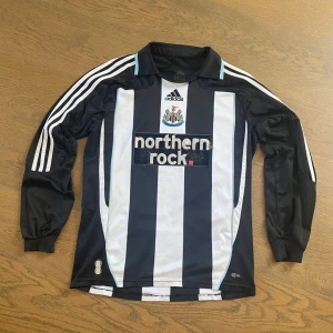 Newcastle United långärmad matchtröja - Säljer en klassisk Newcastle United långärmad fotbollströja från Adidas med svarta och vita ränder, klubbmärke på bröstet och Northern Rock som sponsor. Nummer 14 och N'Zogbia på ryggen. Tröjan har krage och ikoniska Adidas-ränder på ärmarna. Tillverkad i polyester.