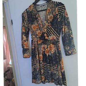 Blommig omlottklänning med långa ärmar - Supersnygg kort omlottklänning från Boohoo med blommigt och randigt mönster i mörka toner. Klänningen har v-ringning, långa ärmar och markerad midja. Perfekt för dig som gillar unika prints och vill sticka ut lite extra.