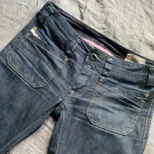 Mörkblå Diesel bootcut jeans - Säljer dessa supersnygga as lågmidjade jeans från diesel! De är bootcut med unika fickor både fram och bak. Jeansen är i storlek W27. De har även söta rosa sömmar på insidan. Bara att fråga om fler bilder eller så om du är intresserad!