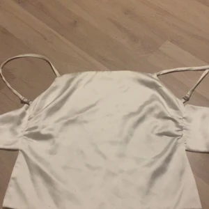 Vit omlott linnetopp med rosett - Säljer en stilren vit linnetopp i satinliknande material med tunna axelband och öppen rygg. Toppen knyts med en stor rosett bak och har en croppad passform. Perfekt för dig som gillar minimalistisk och trendig stil.