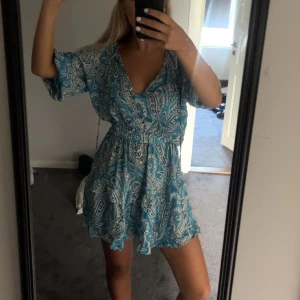 Blå mönstrad kortklänning från Zara - Supersöt blå och vit kortklänning från Zara med paisleymönster, v-ringning med knyt och resår i midjan. Lätt och flowigt material, perfekt för varma dagar. Halvlånga ärmar och volangkant nertill