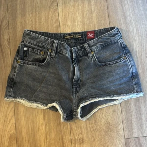 Grå jeansshorts från Superdry - Snygga grå jeansshorts från Superdry med råa kanter och vit spetsdetalj längst ner. Klassisk femficksmodell i tvättad denim, tillverkade av ekologisk bomull. Perfekta för varma dagar och enkel att matcha med allt.
