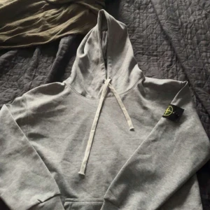 Grå hoodie från Stone Island- inte använd! - Säljer en grå hoodie från Stone Island med klassisk logga på ärmen. Tröjan har huva med snörning och ribbade muddar. Perfekt för dig som gillar streetwear och vill ha en stilren look. Materialet är mjuk bomull som känns skönt mot huden.