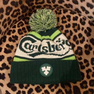 Carlsberg mössa med tofs och logga - Grön, vit och limegrön stickad mössa från Carlsberg med stor tofs på toppen. Mössan har bred uppvikt kant, broderad Carlsberg-logga och en rund patch framtill. Perfekt för dig som vill sticka ut i vinterkylan. Använd 1 gång i nån timme