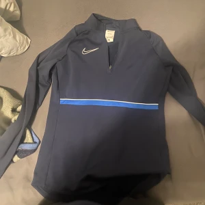 Nike blå långärmad träningströja - Nike Dri-Fit långärmad träningströja i mörkblått med en blå och vit rand över bröstet. Tröjan har half zip och är tillverkad i ett syntetmaterial som andas. Perfekt för dig som gillar sportig stil och vill ha något bekvämt till träningen.
