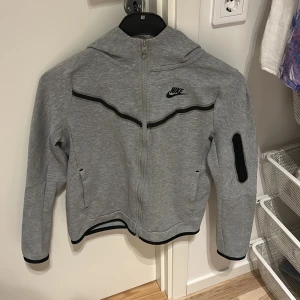 Grå Nike Tech kofta - Grå hoodiejacka från Nike tech med hel dragkedja, svart Nike-logga på bröstet och svarta detaljer längs bröstet och ärmslut. Jackan har huva, lång ärm och en praktisk ficka med dragkedja på ena ärmen. Perfekt för en sportig och chill stil. Använt 4-5 gånger. Värde på 900 kr.