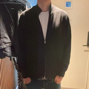 Svart cardigan från Massimo Dutti - Svart cardigan från Massimo Dutti i storlek L. Jackan har en stilren look med dragkedja framtill och är tillverkad i ett mjukt stickat material. Perfekt för dig som gillar en clean och enkel stil med lite edge. Nypris runt 700-800. Skick 7-8/10 (lite nopprig / ser lite använd ut.  tveka inte att höra av dig! 