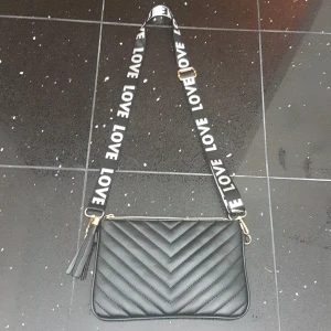 Svart quiltad axelväska med LOVE-band - Snygg svart axelväska i skinnimitation med quiltad framsida och guldfärgade detaljer. Väskan har en bred axelrem med texten LOVE i vitt, dragkedja upptill och en dekorativ tofs. Perfekt storlek för det viktigaste och ger en cool vibe till din outfit. PRIS KAN DISKUTERAS!