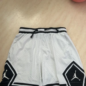 Vita Jordan basketshorts med snörning - Snygga vita Jordan basketshorts med svarta detaljer och logga på benen. Shortsen har elastisk midja med snörning och är tillverkade i luftig mesh för bästa komfort. Perfekta för basket eller träning. Klassisk sportig look med svartvit kontrast.