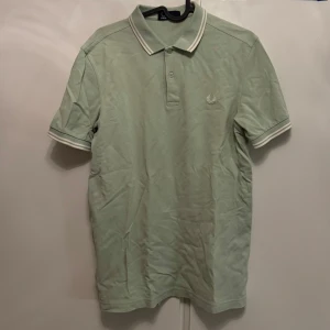 Mintgrön Fred Perry pikétröja - Snygg mintgrön piké från Fred Perry med klassisk krage och vita ränder på krage och ärmslut. Broderad logga på bröstet. Perfekt för dig som gillar stilren och sportig vibe.