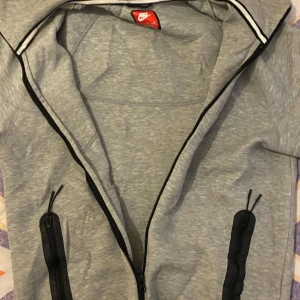 Grå hoodiejacka från Nike med dragkedja - Snygg grå hoodiejacka från Nike med hel dragkedja framtill och svarta detaljer. Jackan har två svarta fickor med dragkedja och en liten Nike-logga på bröstet. Perfekt för chill eller träning, med mjukt material och huva. Storlek M men passar som S.  Pris förslag är öppen för alla.