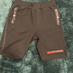 Svarta shorts från Prada - Svarta shorts från Prada med elastisk midja och snygga detaljer i nylon vid fickorna. Stora fickor med tryckknappar och tydlig röd Prada-logga både fram och bak. Tillverkade i 100% bomull, perfekt för en clean och lyxig streetstil. Notera att snören är av och det finns slitage vid snöröjningen samt att knapparna visar tecken på slitningar.
