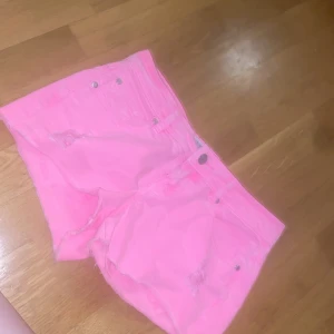 rosa jeansshorts med slitningar - Säljer ett par rosa jeansshorts med råa kanter och slitna detaljer framtill. Shortsen har knapp och dragkedja. Perfekta för sommaren när du vill sticka ut lite extra! Midjemåttet rakt över är ca 38🩷🩷