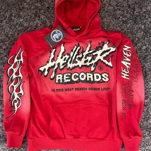 Röd hoodie från Hellstar Records med tryck - Säljer en röd hoodie från Hellstar med stora grafiska tryck både fram och bak. Framsidan har text och logga, medan baksidan har ett stort motiv av ett ansikte med eldliknande hår. Hoodien har känguruficka och tryck på ärmarna.
