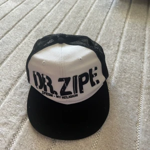 Svartvit keps från Dr. Zipe - Säljer en svartvit keps från Dr. Zipe med stort tryck framtill och texten 'ZIPESIM - MY RELIGION'. Kepsen har svart nät baktill och justerbar snapback-spänne. Perfekt för dig som gillar streetwear och vill sticka ut.