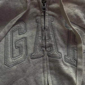 Ljusgrå fleecefodrad hoodie från GAP - Mysig ljusgrå hoodie från GAP med dragkedja och stor broderad logga framtill. Fodrad med vit teddyfleece i huvan för extra värme. Justerbar huva med snören och två fickor framtill.