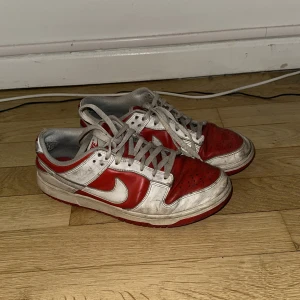 Nike Dunk Low röd och vit - Säljer ett par Nike Dunk Low i rött och vitt med klassisk swoosh på sidan. Skorna har snörning och platt sula, perfekt för dig som gillar streetwear och sneakers med retrostil.