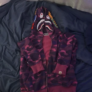 Lila camo hoodie från A Bathing Ape - Säljer en ikonisk lila camo hoodie från A Bathing Ape med hajmotiv på huvan. Tröjan har dragkedja, långa ärmar och ribbade muddar. Perfekt statement-plagg för streetwear-stilen.