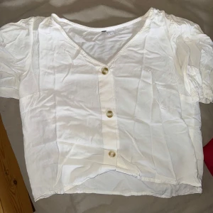Vit blus med puffärm och knappar - Säljer en söt vit blus med v-ringning och tre stora knappar framtill. Blusen har korta puffärmar med resår och är tillverkad i ett tunt, lätt bomullstyg. Perfekt för varma dagar och enkel att matcha. Står att den är storlek 38, men passar mer till 36, S. 