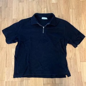 Svart pikétröja med dragkedja från Weekday - Säljer en svart pikétröja från Weekday i storlek L. Tröjan har korta ärmar, klassisk krage och en snygg dragkedja framtill. Perfekt för en stilren och avslappnad look. Ribbat material