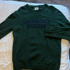 Mörkgrön stickad tröja från Aeronautica Militare - Mörkgrön stickad tröja från Aeronautica Militare med broderad logotyp och text på bröstet. Rund halsringning och ribbade muddar vid ärmslut och nederkant. Perfekt för kyligare dagar.