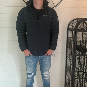 Svart dunjacka från Pelle P - Svart dunjacka från Pelle P med huva och diskret logga på ärmen. Jackan har quiltad design och dragkedja framtill. Perfekt för kyligare dagar och har en stilren look som passar till det mesta.| tveka inte på att höra av er vid intresse 😊|