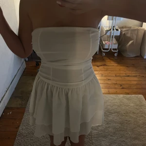 Vit axelbandslös kortklänning med volang - Säljer en söt vit axelbandslös klänning med volangkjol och markerad midja. Klänningen har en luftig och lätt känsla, perfekt för varma dagar eller speciella tillfällen. Stilren och enkel design som är lätt att matcha med accessoarer.