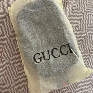 Gucci axelväska med monogram och randigt band - Säljer en snygg axelväska från Gucci i svart med klassiskt GG-monogram. Väskan har en silverfärgad dragkedja framtill och ett justerbart axelband i blått och rött. Perfekt för att bära det viktigaste med stil.