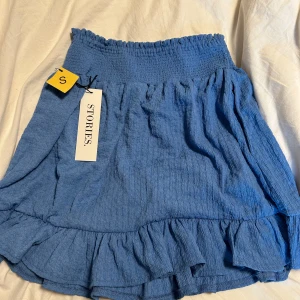 Blå volangkjol  - Supersöt blå kjol med volangdetalj nertill och smock i midjan. Kjolen är kort och har en luftig känsla, perfekt för varmare dagar. Enkel att matcha med olika toppar för en trendig look.