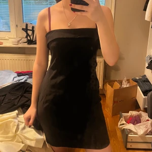 Svart bandouklänning - Säljer en svart kort bandouklänning.  Klänningen har en enkel och stilren design, perfekt för dig som gillar minimalistiskt mode. Dragkedja i ryggen för enkel på- och avtagning.