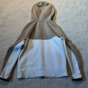 Nike hoodie i beige och vitt - Storlek 158-170 cm junior. Säljer en snygg hoodie från Nike i beige och vitt med svarta detaljer. Hoodien  har huva, dragkedja framtill och två svarta fickor. Perfekt för en sportig och avslappnad stil.