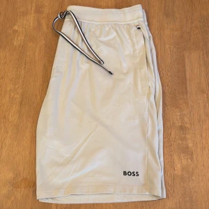 Beige shorts från BOSS med snörning - Snygga beige shorts från BOSS med svartvit snörning i midjan och diskret logga på benet. Shortsen har sidofickor och är tillverkade i ett mjukt, lätt material som passar perfekt för varma dagar.
