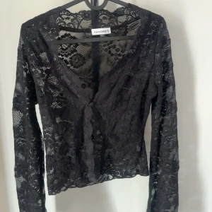 Svart spetsig croppad blus från Venderby - Säljer en svart långärmad blus från Venderby i genombruten spets med v-ringning och croppad passform. Blusen har dekorativa kanter och knäppning framtill, perfekt för en trendig look. Storlek M/L men passar en S/M