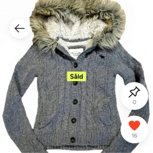 Abercrombie & Fitch stickad hoodie med fuskpäls - Mysig grå stickad hoodie från Abercrombie & Fitch med knappar framtill och stor huva fodrad med mjuk fuskpäls. Kabelstickat mönster och ribbade muddar. Perfekt för kyliga dagar.