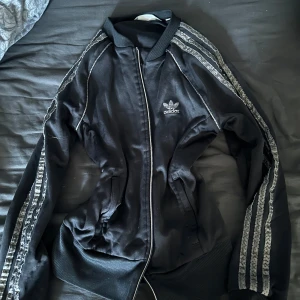 Svart Adidas bomberjacka med dragkedja - Svart bomberjacka från Adidas med klassiska vita ränder längs ärmarna och dragkedja framtill. Jackan har ribbade muddar vid ärmslut och midja samt Adidas-logga på bröstet. Perfekt för en sportig och avslappnad stil.