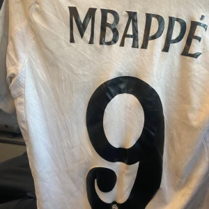 Real Madrid Mbappé 9 fotbollströja - Säljer en vit Real Madrid fotbollströja med Mbappé och nummer 9 på ryggen. Tröjan har svart tryck, klubbmärke på bröstet och Adidas-logga. Emirates Fly Better som sponsor framtill. Perfekt för fans!