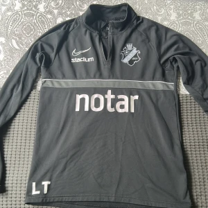 Svart AIK Nike träningströja med dragkedja - Svart långärmad träningströja från Nike med AIK:s klubbmärke, och sponsorer. Tillverkad i Dri-Fit material som andas. Perfekt för fotbollsträning eller sportiga dagar.