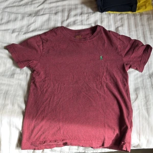 Vinröd t-shirt från Polo Ralph Lauren - Vinröd t-shirt från Polo Ralph Lauren med klassisk passform och liten grön broderad logga på bröstet. Tillverkad i mjuk bomull och har korta ärmar. Perfekt för en avslappnad stil.