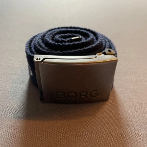 Mörkblått tygbälte från Björn Borg - Säljer ett mörkblått tygbälte från Björn Borg med silverfärgat spänne där det står 'BORG'. Bältet har tydlig logga längs hela bandet och passar perfekt till jeans eller casual outfits.