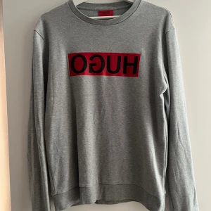 Grå sweatshirt från Hugo Boss - Säljer en grå sweatshirt från Hugo Boss med röd och svart logga tryckt på bröstet. Tröjan har rund halsringning och långa ärmar. Perfekt för en avslappnad och stilren look.