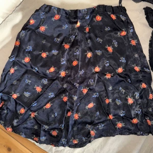 Svart blommig satinkjol - Säljer en svart satinkjol med små röda och blå blommor. Kjolen har en kort längd och en lätt glansig yta som ger en snygg look. Perfekt att matcha med en enkel topp för en trendig outfit. Är lite högmidjad men kan dras ner och då bli lite längre i längden. Dragkedja på sidan.