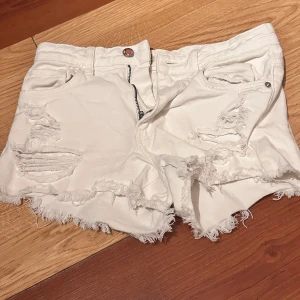 Vita jeansshorts med fransar från Tally Weijl - Säljer ett par vita jeansshorts från Tally Weijl med slitna detaljer och fransig kant. Shortsen har klassisk femficksmodell, knapp och dragkedja framtill. Perfekta för sommaren och ger en cool, avslappnad look.