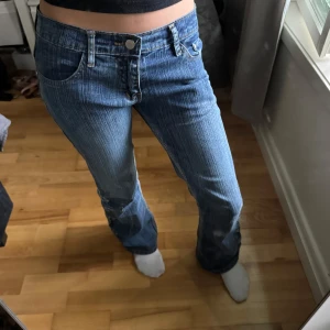 Blå jeansbyxor med detaljer från Army Jeans - Säljer ett par blå lowwaist bootcut jeans från Army Jeans med unika knappliknande detaljer längs utsidan av benen. Byxorna har normal passform, klassisk femficksdesign och snygg tvättad look. Perfekta för dig som vill sticka ut med din denimstil. Jag säljer dessa eftersom de är lite för stora för mig så därför kommer dom inte till användning!