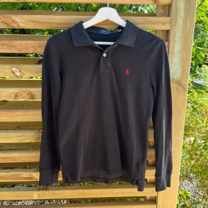 Svart långärmad pikétröja från Polo Ralph Lauren - En svart långärmad pikétröja från Polo Ralph Lauren i storlek XS Slim Fit. Tröjan är köpt i Ralph Lauren egna butik i USA och är därför ÄKTA. Tröjan är nästan helt ny och endast använd ett fåtal gånger. Den har väldigt bra och smal passform. Tröjan har ett påsytt rött emblem på vänster bröst. 