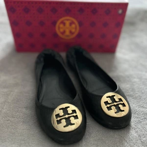 Svarta ballerinaskor från Tory Burch - Säljer ett par klassiska svarta ballerinaskor från Tory Burch med stor guldfärgad logga framtill. Skorna har rund tå och platt sula, perfekta för en stilren look. Kommer med originalkartong och dustbag. Endast använda fåtal gånger.