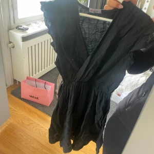 Svart omlottklänning med volang - Supersöt svart kortklänning med omlott och volangdetaljer vid ärmarna. Klänningen har v-ringning och är ärmlös, perfekt för en trendig look. Tillverkad i ett lätt tyg som känns skönt mot huden.