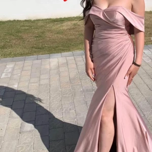 Ljusrosa offshoulder långklänning med slits - Elegant ljusrosa långklänning med offshoulder och draperad överdel. Klänningen har en markerad midja, figurnära passform och hög slits framtill. Perfekt för bal eller fest. 