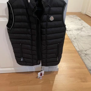 Svart dunväst från Moncler - Snygg svart dunväst från Moncler med diskret logotyp på bröstet. Västen har dragkedja framtill och två fickor med dragkedja. Perfekt för lager-på-lager och har en sportig, modern look.